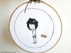 an embroidery hoop with a black and white drawing of a girl on it, next to two buttons