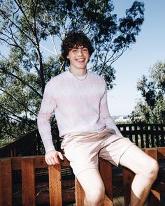 a young man sitting on a wooden bench wearing shorts and a white sweater with trees in the background