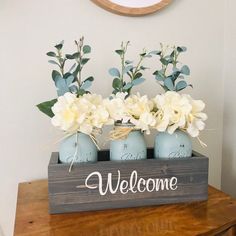 three mason jars with flowers in them are sitting on a wooden box that says welcome