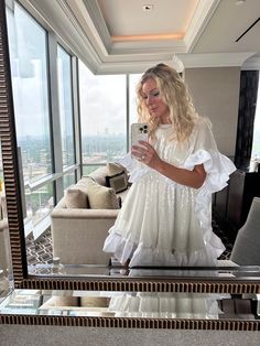 a woman in a white dress taking a selfie with her cell phone while standing in front of a large window