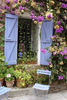 an open window with blue shutters and purple flowers on the wall next to two chairs