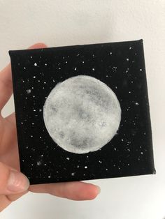 a hand holding up a small black and white painting with the moon in the background