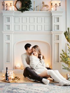 a man and woman sitting on the floor in front of a fireplace with christmas decorations