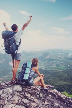 two people standing on top of a mountain with backpacks and pointing at the sky