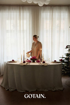 A Christmas-themed dining room with a festive table setting, a glowing Christmas tree, and sheer linen curtains in sand filtering soft natural light.