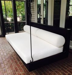 a white bed sitting in front of a window on top of a brick floor next to a black framed mirror