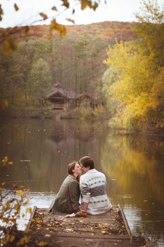 two people sitting on a dock kissing in front of a body of water with trees around them