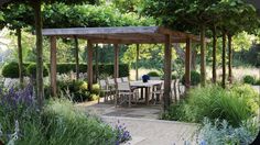 a table and chairs under a pergoline surrounded by trees in the middle of a garden