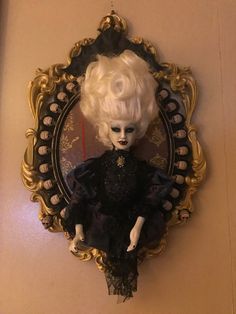 a doll with white hair and black dress sitting in front of a gold framed mirror