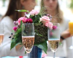 two champagne flutes are sitting on a table with pink flowers in the background and a woman standing behind them