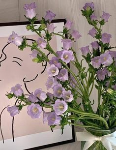 a vase filled with purple flowers sitting next to a framed drawing on a wall behind it