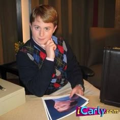 a young man sitting at a table with his hand on his chin and looking into the camera