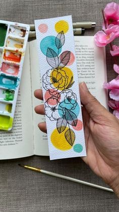 a hand holding an open book with flowers painted on it and paintbrushes next to it