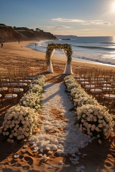 An elegant beach wedding setup features a floral aisle made of white petals leading to an arch adorned with white flowers. Wooden chairs flank the aisle, and the scene is set against a backdrop of the ocean and cliffs under a sunset sky. Simple Wedding Beach Ideas, Small Beach Wedding Bridesmaid, Micro Wedding Ideas Beach, Magical Beach Wedding, Old Money Beach Wedding, Wedding Venue Ideas Beach, Wedding Decorations Beach, Outdoor Wedding Beach, Beach Marriage