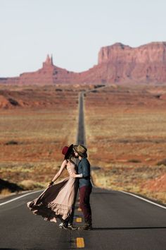 a man and woman standing on the side of an empty road with mountains in the background