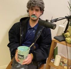 a man sitting on a chair holding a cup with something painted on his face in front of him