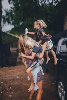 a woman holding a child in front of a black truck with trees and dirt on the ground