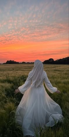 a woman in a white dress is walking through the grass at sunset with her arms outstretched