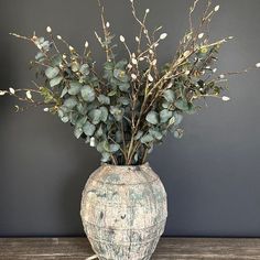 a vase filled with lots of green plants on top of a wooden table next to a gray wall