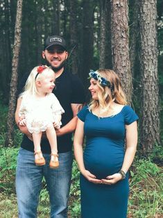 a man and woman holding a baby standing in the woods with their pregnant child, who is wearing a blue dress