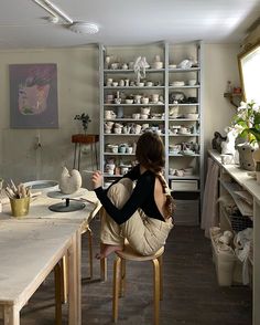 a woman sitting at a table in a room with shelves and pots on the wall