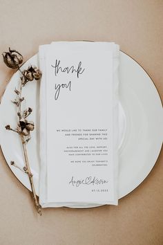 a white plate topped with a piece of paper next to a dried flower on top of a table