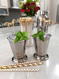 two silver cups with mint leaves and straws on a marble counter top in front of a vase