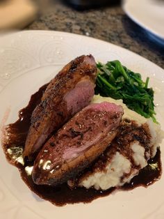 some meat and mashed potatoes on a white plate with greens in the middle, sitting on a marble table