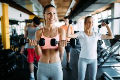 two women lifting dumbbells in a gym while another woman watches from the sidelines