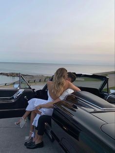 a man and woman sitting on the back of a convertible car next to an ocean