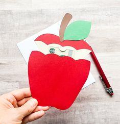 someone is holding an apple cut out from paper with a pencil on the table next to it