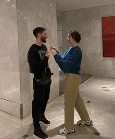 two people standing in a room talking to each other and smiling at one another with their hands together