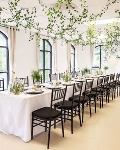 a long table set up with black chairs and greenery hanging from the ceiling in front of large windows