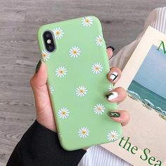 a woman holding a green phone case with white daisies on it and the ocean in the background