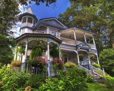 a large blue and white house in the woods with flowers on it's front porch