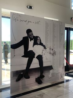 a man and woman sitting on top of a couch in front of a large poster