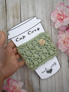 someone is holding up a cup cozy in front of some pink flowers on a wooden table