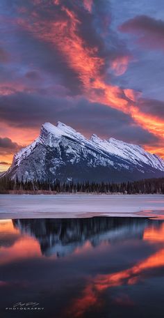 the mountains are covered in snow as the sun goes down over water and trees is reflected in the lake's surface