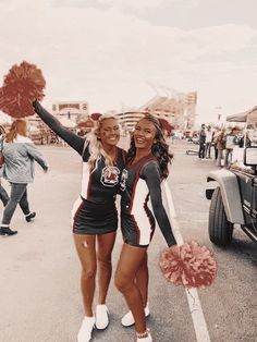 two cheerleaders standing in the parking lot holding pom poms and smiling