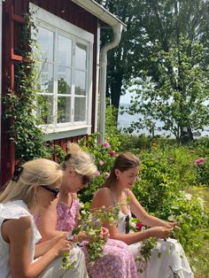 three women sitting on a bench in front of a house with flowers around their ankles