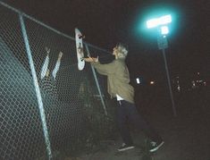 a man holding a skateboard next to a chain link fence in the dark at night