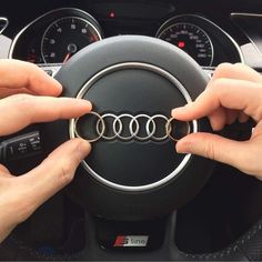 two hands on the steering wheel of a car holding onto an audi emblem with gold rings