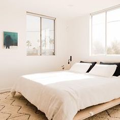 a white bed sitting in a bedroom next to two large windows and a rug on the floor