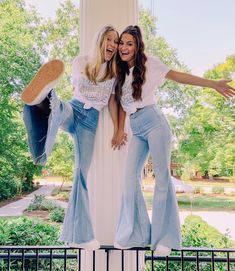 two women are posing for the camera in front of a white pillar with trees and bushes behind them