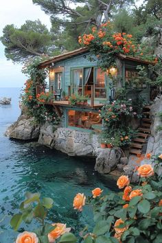 the house is surrounded by flowers and greenery on the cliff above the water's edge