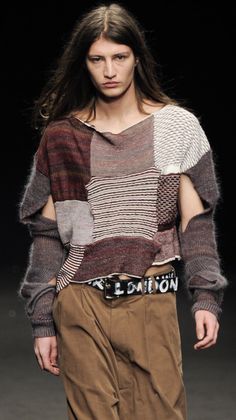 a man with long hair wearing a multicolored sweater and brown pants on the runway