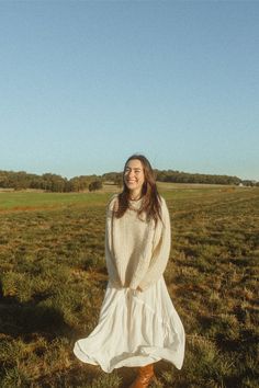 a woman standing in the middle of a field wearing a white dress and brown boots