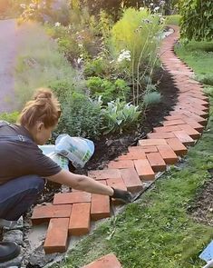 a woman laying bricks on the ground in front of some plants and flowers near a brick path