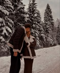 a man and woman standing in the snow near pine trees with their noses close to each other