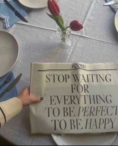 a person is holding up a newspaper on a table with plates and flowers in the background